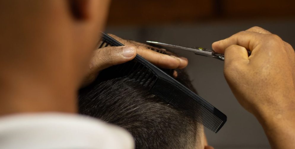 A male barber in a salon trims curtain bangs on a man, with the fringe parted in the middle and falling softly around th